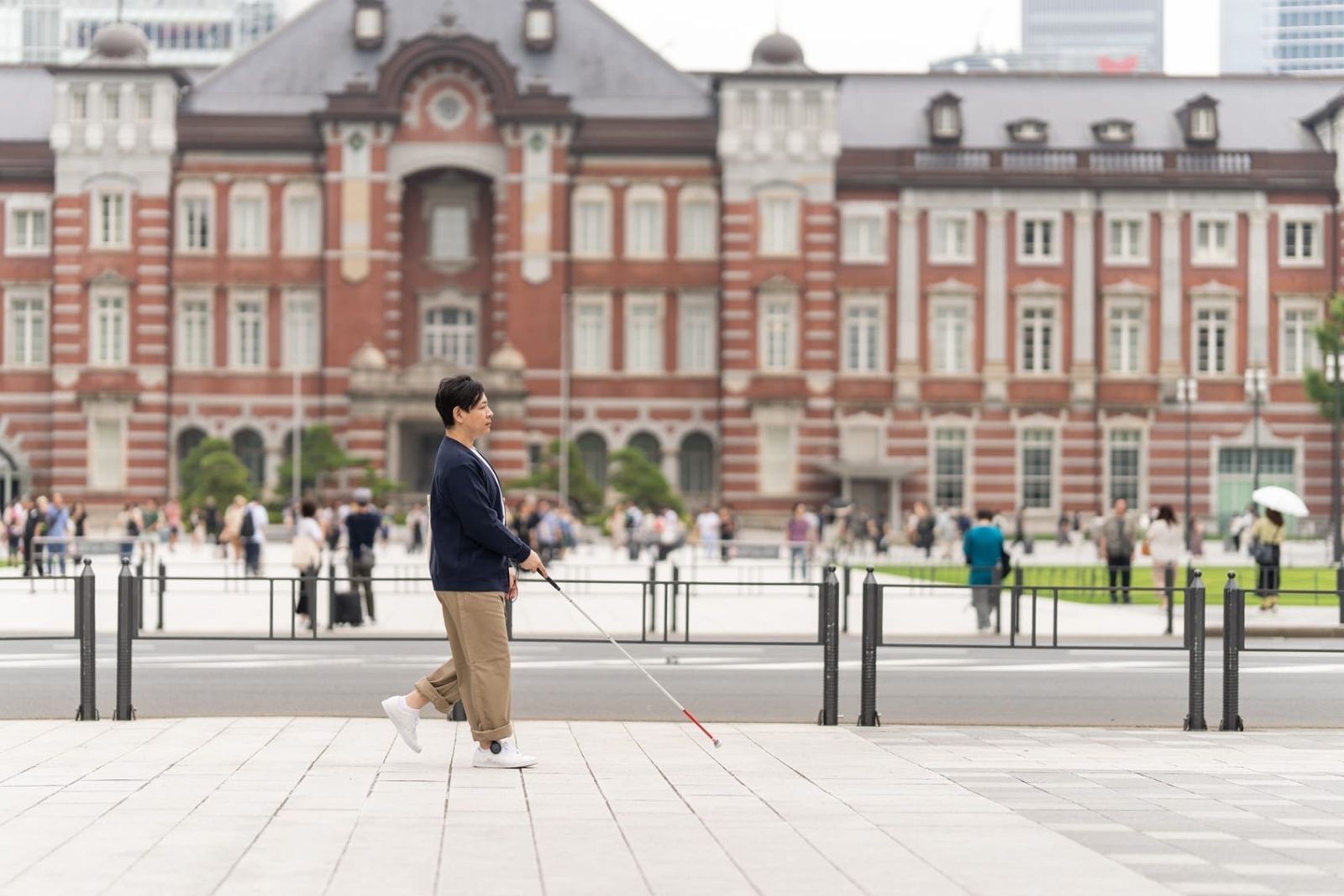 白杖を使って都市空間を歩く人物の全身と、その周囲の街並みが写った写真