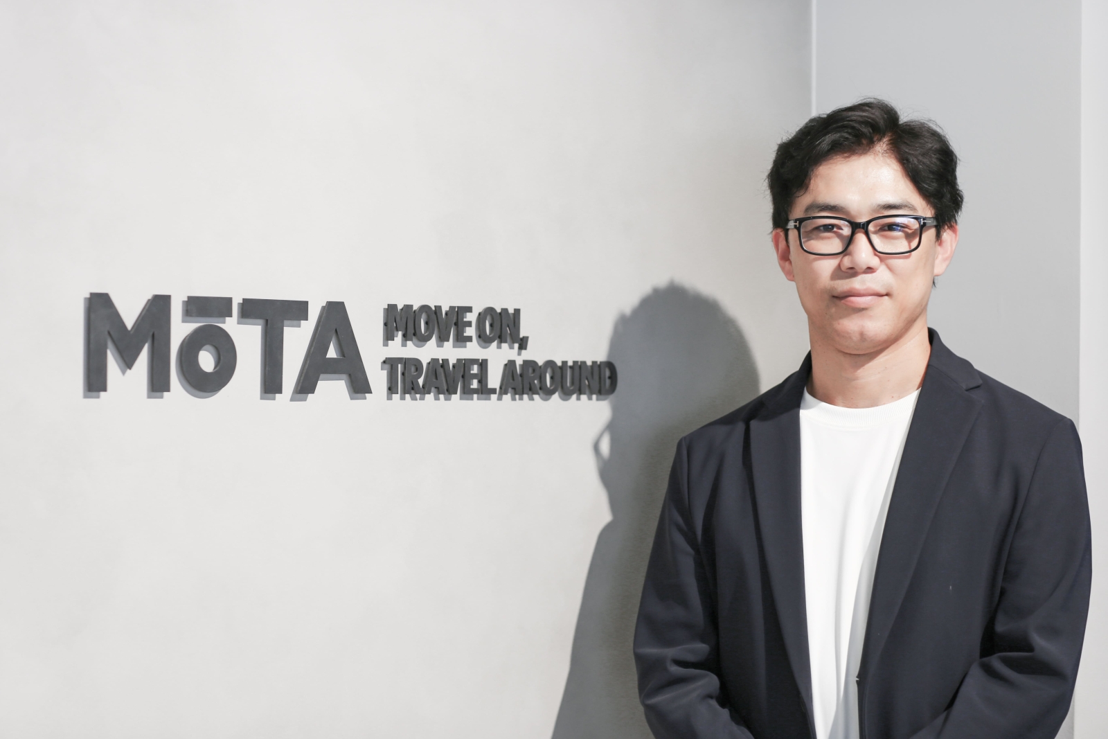 株式会社MOTAのロゴサインの前に立つ、代表取締役社長 佐藤大輔氏